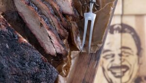 A smoked brisket rests on a wooden cutting board, with slices arranged on parchment paper beside a meat fork. The background features the partially visible logo of Hoots Smokehouse, a Black-owned BBQ restaurant known for its expertly crafted smoked meats.