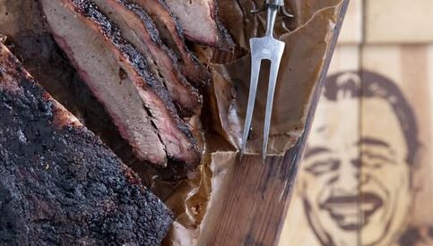 A smoked brisket rests on a wooden cutting board, with slices arranged on parchment paper beside a meat fork. The background features the partially visible logo of Hoots Smokehouse, a Black-owned BBQ restaurant known for its expertly crafted smoked meats.